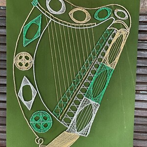 May include: A green, white, and gold string art piece depicting a harp. The harp is made of string wrapped around nails hammered into a wooden board.