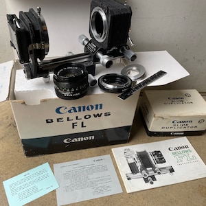 May include: A Canon Bellows FL camera attachment with a black bellows, a lens, and a slide duplicator. The camera attachment is on a white surface with a box labeled "Canon Bellows FL" and a box labeled "Canon Slide Duplicator".