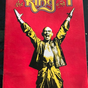 May include: A red poster with the text "the King and I" in yellow lettering. A man in a yellow and black outfit is standing with his arms raised in the air.