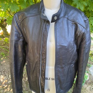 May include: A black leather jacket featuring a mandarin collar with a snap button closure, a full-length silver-tone metal front zipper, and long sleeves. The jacket has a smooth texture and is displayed on a dress form with visible stitching.