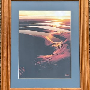 May include: Framed print of a landscape photograph showing a river winding through a desert at sunset. The sky is a vibrant orange and pink, and the river is a deep blue. The artist's signature, "Kimo", is in the bottom right corner.