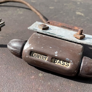 May include: Vintage brown "Country Bass" device with a metal plate and screws attached. A plug with multiple prongs and a wire are connected to the device. The device has a rounded shape with the words "COUNTRY BASS" in a rectangular window.