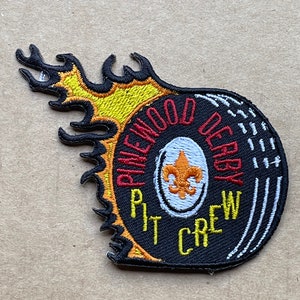 May include: Black and yellow embroidered patch with a tire design. The tire has the text "Pinewood Derby Pit Crew" and a fleur-de-lis in the center.