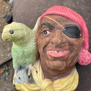 May include: A ceramic figurine of a pirate with a red hat, an eyepatch, and a yellow scarf. A green parrot is perched on the pirate's shoulder.