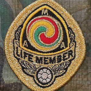 May include: Gold embroidered patch with a black and gold swirl design in the center. The swirl is red, green, and yellow. The text "LIFE MEMBER" is below the swirl. A black and white soccer ball with wings is below the text.