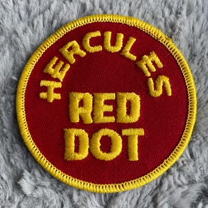 May include: A red embroidered patch with yellow stitching. The patch is circular and features the text "Hercules Red Dot" in yellow.