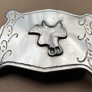May include: Silver belt buckle with a detailed engraved design of a saddle. The buckle is rectangular with rounded corners and has a decorative border.