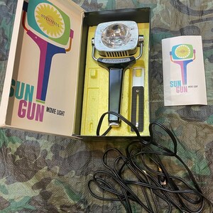 May include: A vintage Sylvania Sun Gun movie light in its original box. The light is black with a silver reflector and a black cord. The box is white with a yellow and blue illustration of the light.