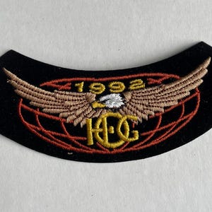 May include: Black felt patch with red and yellow embroidered design. The design features a globe with an eagle with outstretched wings and the text "1992 H.O.G."