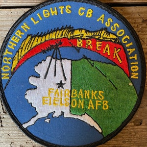 May include: A blue, round patch with a black border. The patch features a yellow, red, and white aurora borealis design with the text "NORTHERN LIGHTS CB ASSOCIATION BREAK" in yellow. The bottom of the patch shows a white and green outline of Alaska with the text "FAIRBANKS Eielson AFB" in yellow.