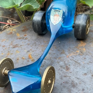 May include: A translucent blue toy vehicle with a long, slender body and a pointed front section. It features two large black rear wheels and two smaller gold-colored front wheels with a perforated design. A shield-shaped emblem with an eagle is visible.