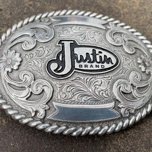 May include: Silver belt buckle with an ornate design and the words "Justin Brand" in black lettering.