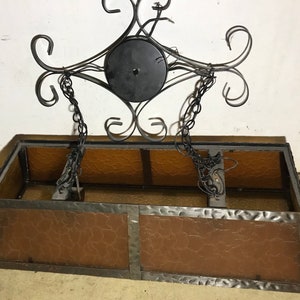 May include: A rectangular, brown glass light fixture with a wrought iron frame. The fixture has a chain hanging from it and a black metal plate at the top.