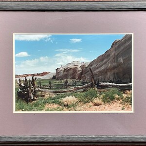 May include: A framed landscape photograph featuring a desert scene with a wooden fence in the foreground. The image captures a blue sky with clouds, a rock formation, and green vegetation. The frame is gray.
