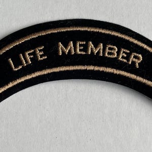 May include: Black and gold embroidered patch with the text "LIFE MEMBER" in a curved shape.