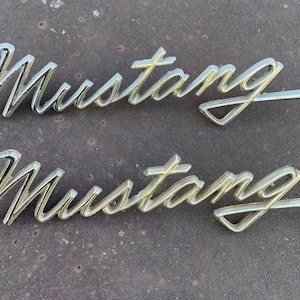 May include: Two silver-colored metal "Mustang" emblems. The emblems are in a cursive font and have a slightly raised design.