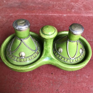 May include: A green ceramic salt and pepper set with silver-colored metal accents. The set includes two shakers and a central handle, all resting on a matching tray. The shakers have a bulbous shape and are adorned with decorative metalwork.