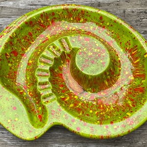 May include: A green ceramic ashtray with red and yellow splatter paint. The ashtray has a deep center well and a wide, shallow outer ring.
