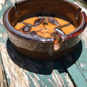 May include: A brown ceramic ashtray with a dark brown glaze and a decorative design. The ashtray has a round shape with a small indentation for cigarettes.