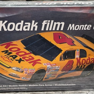 May include: A model kit box for a 1:25 scale model of a yellow Kodak film Monte Carlo race car. The box features a photo of the car with the Kodak Max film logo and the number 4 on the side.