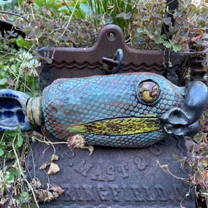 May include: A ceramic fish sculpture with a textured blue and green scale pattern, a yellow-speckled side, and a prominent eye. The fish has a dark blue tail fin and a dark, rounded snout. It is positioned horizontally on a dark metal object.