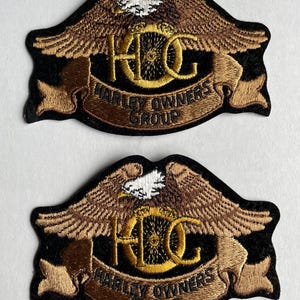 May include: Two brown and black embroidered Harley Owners Group (HOG) patches with a gold eagle and the letters HOG in a circle. The patches have a brown banner with the words Harley Owners Group.