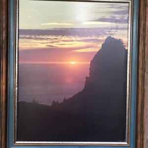 May include: Framed print of a sunset over the ocean with a large rock formation in the foreground. The sky is a vibrant pink and orange, and the water is a deep blue. The print is in a wooden frame with a blue and silver border.