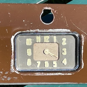 May include: A brown metal dashboard panel with a circular gauge featuring black numbers 0 through 12 and a silver needle. The gauge is surrounded by a silver frame and has a small hole in the center.