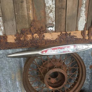 May include: A rusty metal wheel with a chrome Chevrolet 6400 emblem on top. The wheel is on a wooden background with a rusty metal cutout of a western scene.