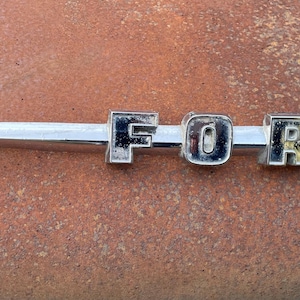 May include: A vintage chrome "FORD" emblem, featuring the brand name in capital letters. The emblem is mounted on a silver bar with a pointed end. The background is a rusty, textured surface.