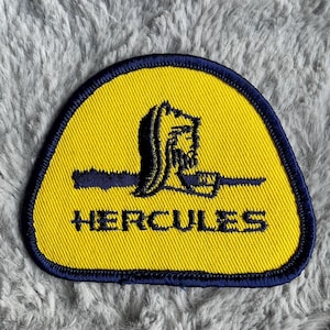 May include: A yellow fabric patch with a blue border. The patch features a black silhouette of a man lifting a weight with the word "HERCULES" in black text below.