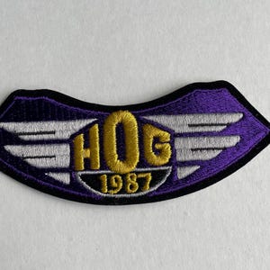 May include: Purple and white embroidered patch with the text "HOG 1987" and wings.