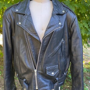 May include: A black leather motorcycle jacket featuring a wide notched collar, silver-toned metal zippers, and a front belt with a buckle. The jacket includes a diagonal chest zipper pocket, a small flap pocket with a snap button, and shoulder epaulets.
