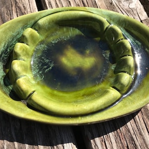 May include: A green ceramic ashtray with a raised, wavy rim and four indentations for cigarettes. The ashtray has a mottled green glaze with darker green and black accents.