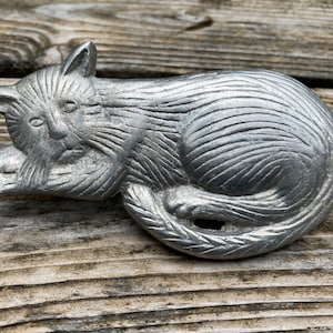 May include: A silver metal figurine of a cat lying down with its head resting on its paws. The cat has a detailed, textured fur pattern.