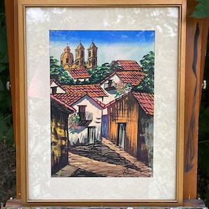 May include: A framed watercolor painting depicting a colorful, sun-drenched Mexican village with red-tiled roofs, whitewashed walls, and a church with twin bell towers in the background.
