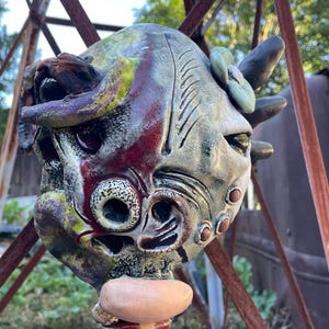 May include: A ceramic mask featuring a multi-colored glaze in shades of green, red, and grey. The mask has an asymmetrical, textured surface with protruding elements, including a smooth, light-colored nose and a small flower-like detail.