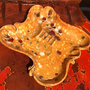 May include: A vintage ceramic ashtray with a unique, abstract design. The ashtray is brown with red and white speckles and has three indentations for cigarettes.