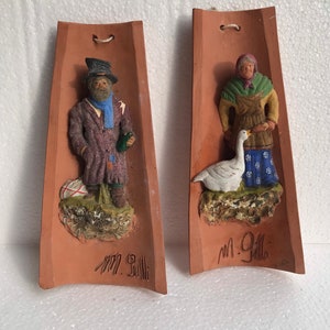 May include: Two terracotta wall plaques with raised figures of a man and a woman. The man is wearing a brown coat, a blue scarf, and a black hat. He is holding a bottle in his right hand. The woman is wearing a green apron, a brown shawl, and a white head covering. She is holding a white goose in her left hand. Both plaques are signed "M. Gilla".
