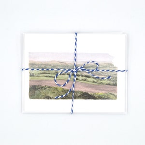 May include: A set of blank greeting cards tied together with blue and white twine. The cards feature a watercolour painting of a landscape with rolling hills and a field.