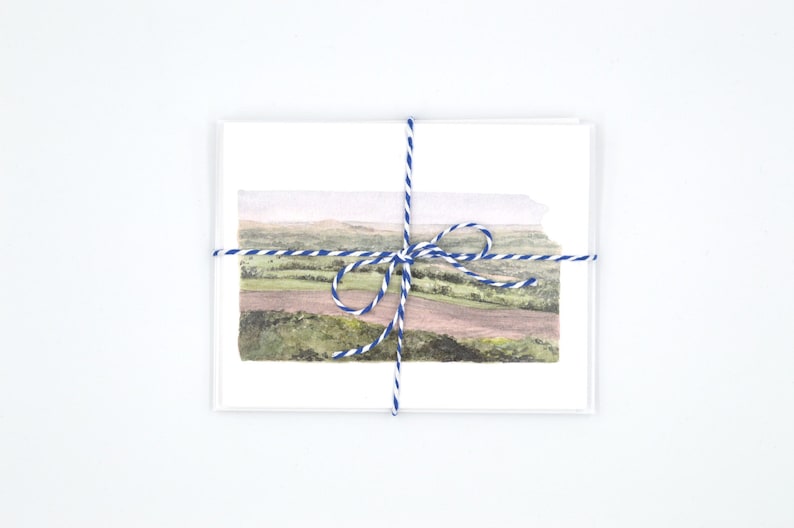 May include: A set of blank greeting cards tied together with blue and white twine. The cards feature a watercolour painting of a landscape with rolling hills and a field.