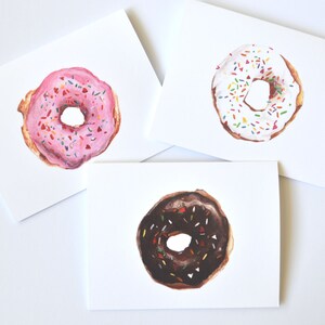 May include: Three blank greeting cards with illustrated donuts. The donuts are pink with sprinkles, white with sprinkles, and chocolate with sprinkles.