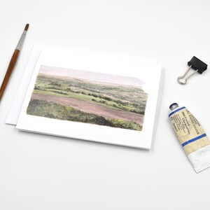 May include: A watercolour painting of a landscape scene on a white card. The painting depicts a field with rolling hills and trees in the distance. The card is blank and ready for a message.
