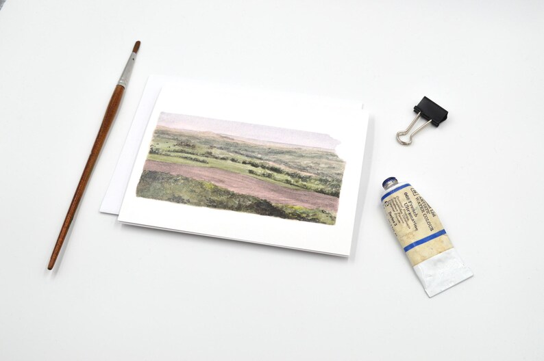 May include: A watercolour painting of a landscape scene on a white card. The painting depicts a field with rolling hills and trees in the distance. The card is blank and ready for a message.