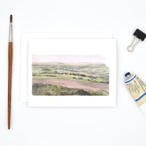 May include: A watercolour painting of a landscape with rolling hills and a field in the foreground. The painting is on a white card.