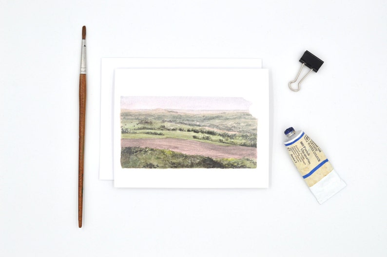 May include: A watercolour painting of a landscape with rolling hills and a field in the foreground. The painting is on a white card.