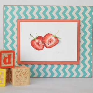 Op de afbeelding: Een ingelijst aquarel schilderij van twee gehalveerde aardbeien op een witte achtergrond. De lijst is turquoise met een wit zigzagpatroon en een koraal gekleurde binnenlijst. Het schilderij is gesigneerd "R. Alvare." Drie houten blokken met de letters "E", "D" en "2" bevinden zich op de voorgrond.