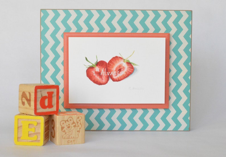 Op de afbeelding: Een ingelijst aquarel schilderij van twee gehalveerde aardbeien op een witte achtergrond. De lijst is turquoise met een wit zigzagpatroon en een koraal gekleurde binnenlijst. Het schilderij is gesigneerd "R. Alvare." Drie houten blokken met de letters "E", "D" en "2" bevinden zich op de voorgrond.