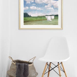 May include: A framed watercolor painting of a field with white laundry hanging on a clothesline. The sky is blue with white clouds. The painting is hanging on a white wall with a white chair and a woven basket in the foreground.