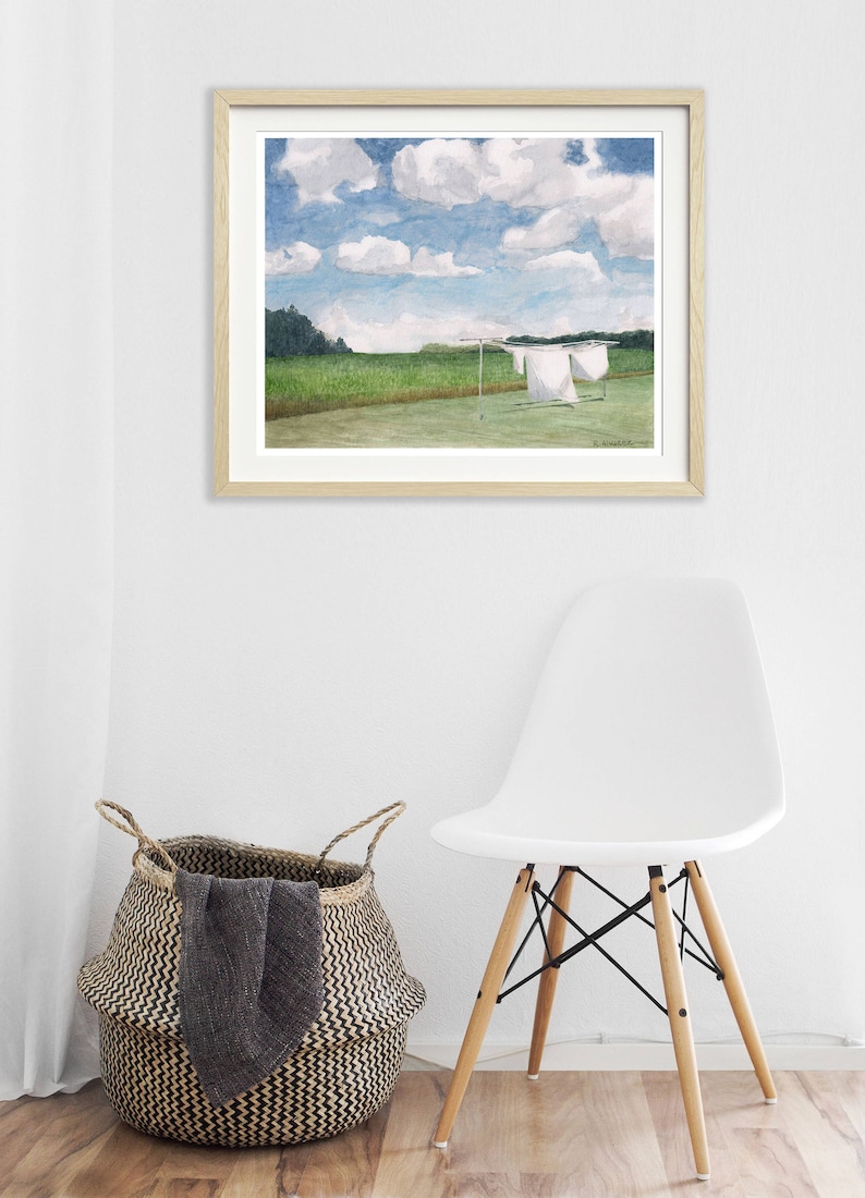 May include: A framed watercolor painting of a field with white laundry hanging on a clothesline. The sky is blue with white clouds. The painting is hanging on a white wall with a white chair and a woven basket in the foreground.
